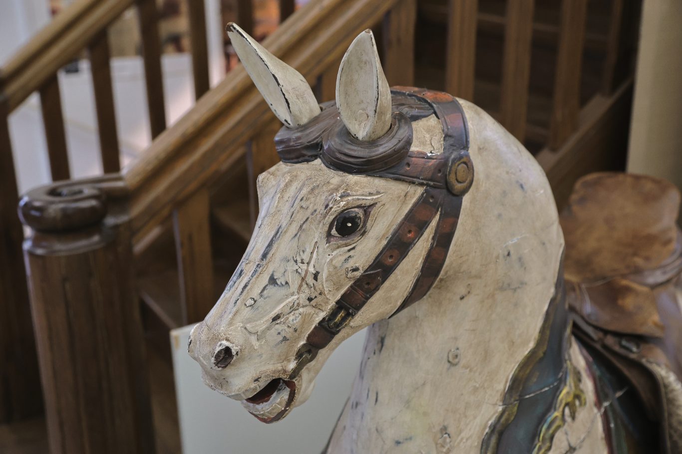 Weißes Holzpferd mit Brille, kunstvoll verziert, vor einer Holztreppe.