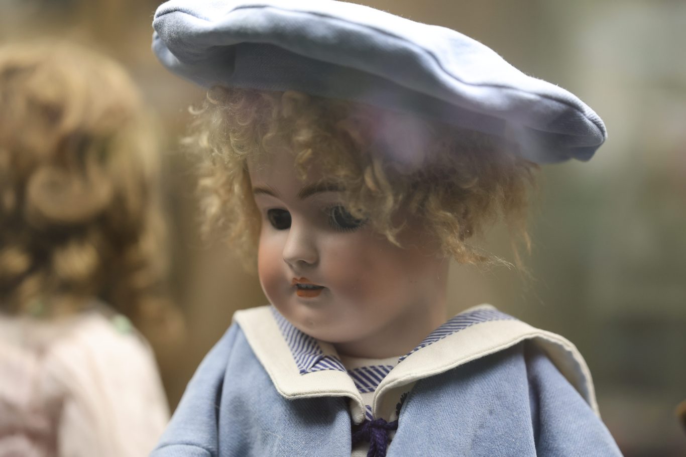 Antike Puppe mit blonden Locken und blauem Hut, in einem Vitrinenausstellungsraum.